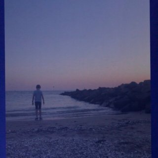 Un enfant sur une plage lors d’un coucher de Soleil, les pieds presque dans l’eau. L’image est bleutée.